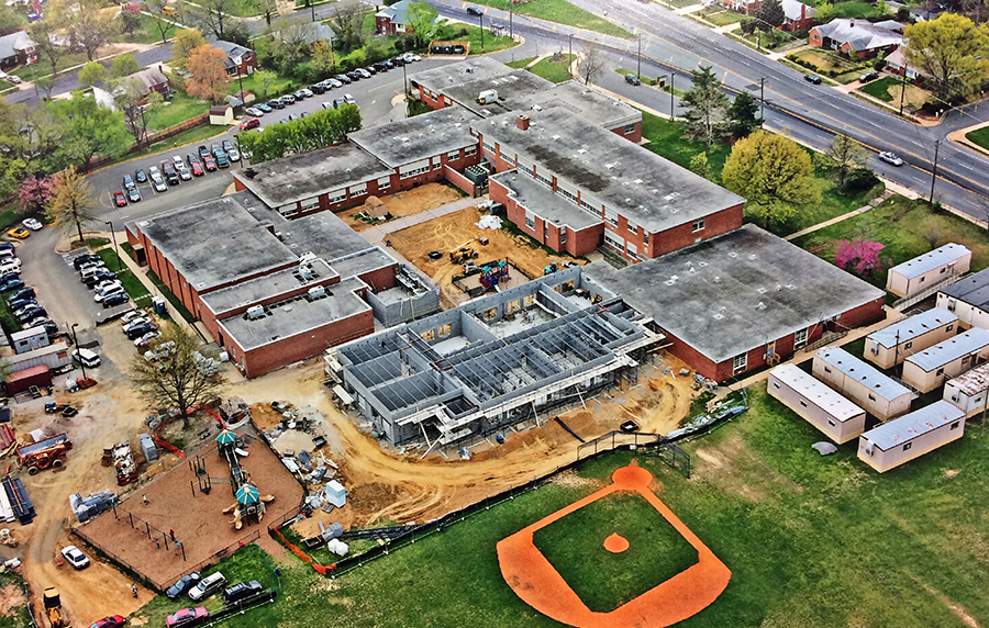 Aerial photograph of Lynbrook Elementary School taken from the northeast. A large cluster of portable classroom trailers is visible on the far right of the screen. A new classroom wing is being constructed on the rear of the building. The grounds around the courtyard and rear of the building have been heavily disturbed by construction. Sections have been fenced off and walkways have been created to guide students safely from the building to the playgrounds. Trees in the interior courtyard have been removed and construction equipment is visible in these areas. The cinderblock walls of the addition are complete, but the addition is not yet under roof.  
