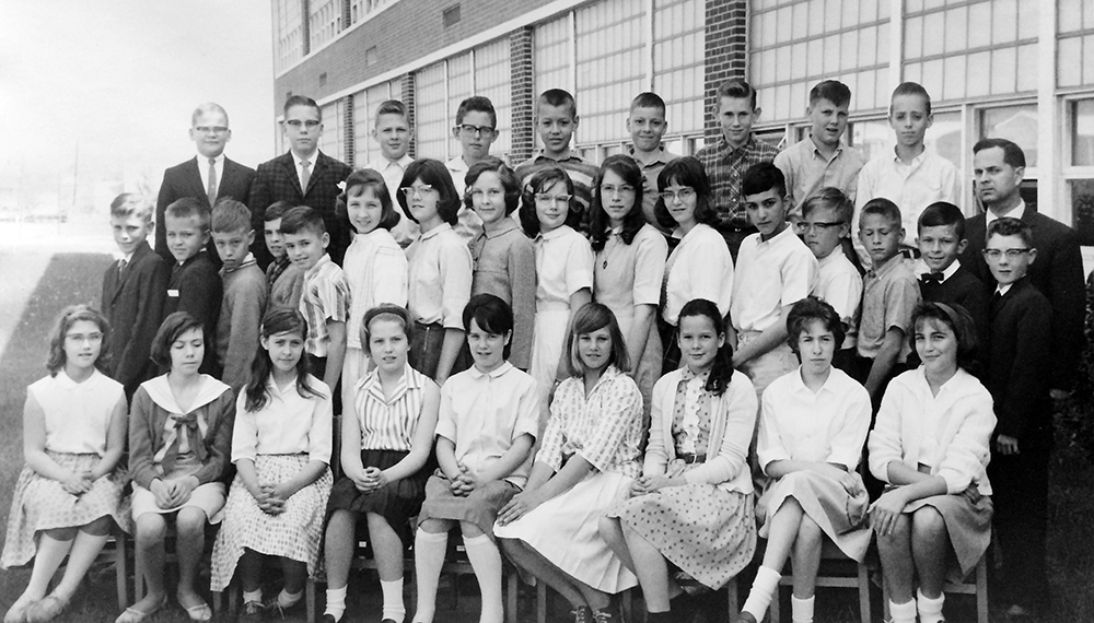 Black and white class photograph taken in the spring of 1964. Mr. Sheridan's class is posed outside with the two-story classroom wing of the building visible behind them. 34 children and their teacher are pictured. They are arranged in three rows with the first row, all girls, seated in chairs. The last row, all boys, are standing on chairs. Mr. Sheridan is visible on the far right.