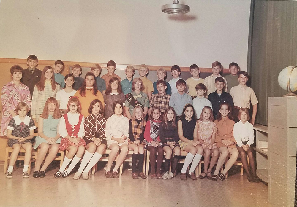 Color class photograph taken during the 1970 to 1971 school year. 34 children and their teacher are posed in a classroom. The children are arranged in three rows in front of a blackboard. Green curtains have been drawn across the windows on the right side of the picture. A circular metallic light fixture is visible hanging down from the ceiling.