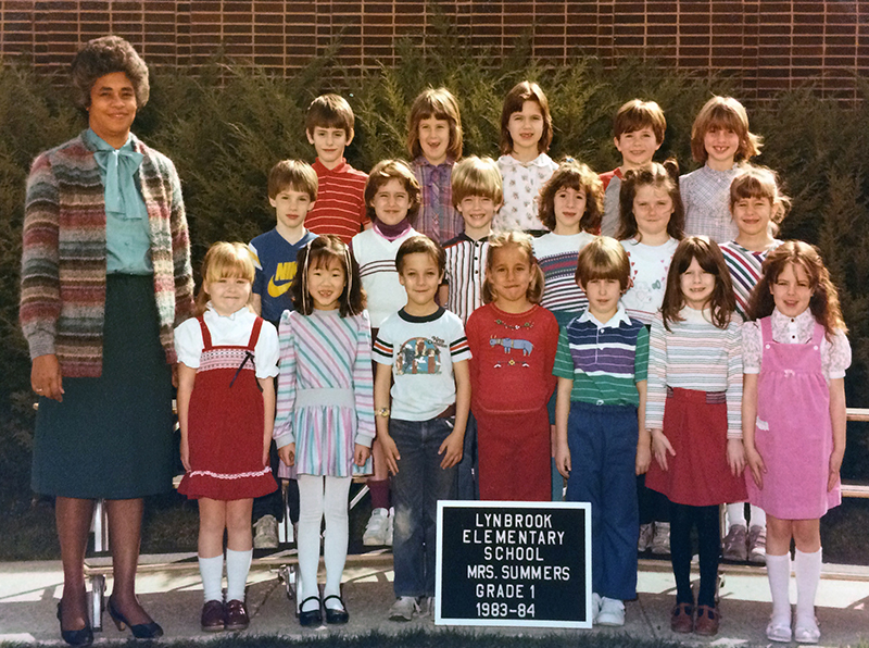 Color photograph showing Mrs. Summers' first grade class. 18 children are pictured. They are standing in three rows on a riser placed outside on the sidewalk next to the cafeteria. Mrs. Summers can be seen standing on the far left of the image.