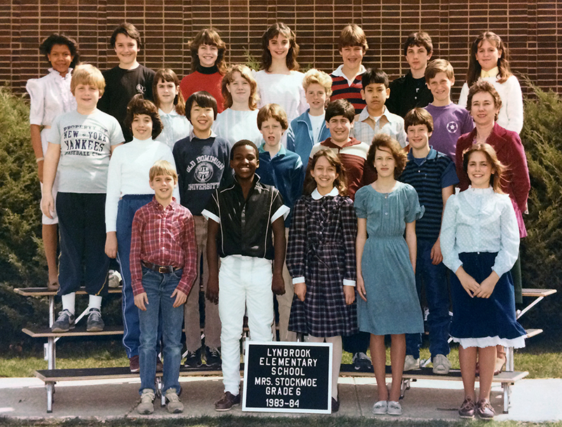 Color photograph of Mrs. Stockmoe's sixth grade class. 23 children are pictured. They are standing in four rows on a riser placed outside on the sidewalk next to the cafeteria. Mrs. Stockmoe is standing on the right in the second row.