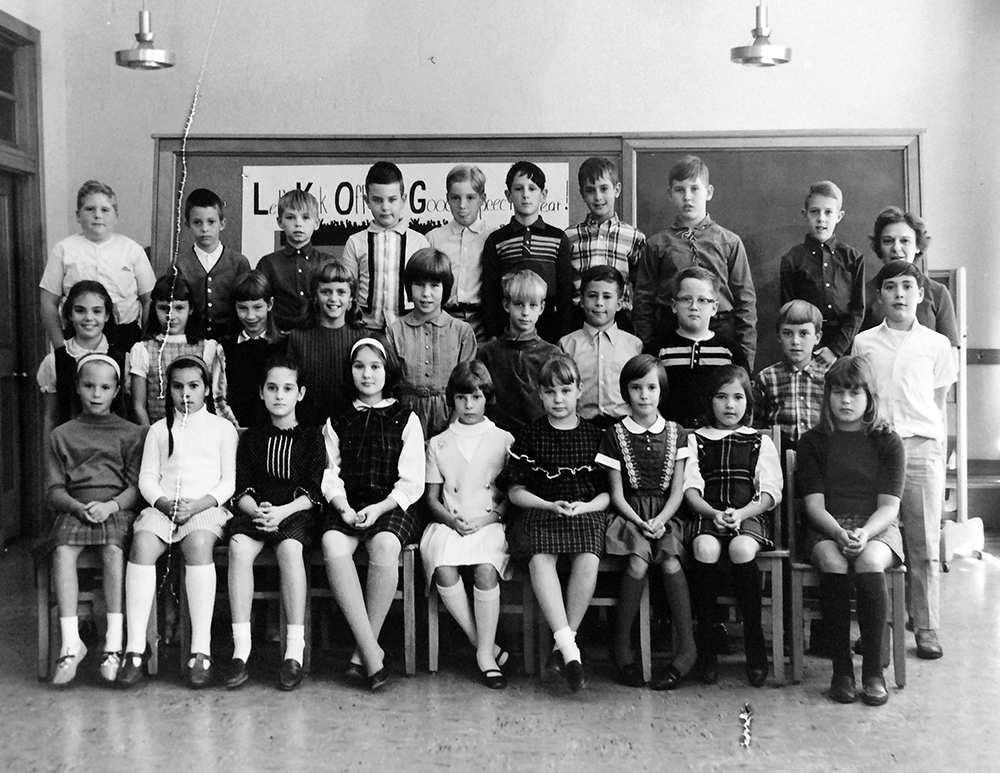 Black and white class photograph taken in October 1966. No name is given on the photo, but a female teacher is visible on the far right of the picture. The photograph was taken in a classroom. 28 children are pictured. The children are arranged in three rows in front of a blackboard and a cork board. The classroom door is visible on the left. It is made out of wood and there is a transom above the doorway. Two circular metallic light fixtures are visible hanging down from the ceiling. 