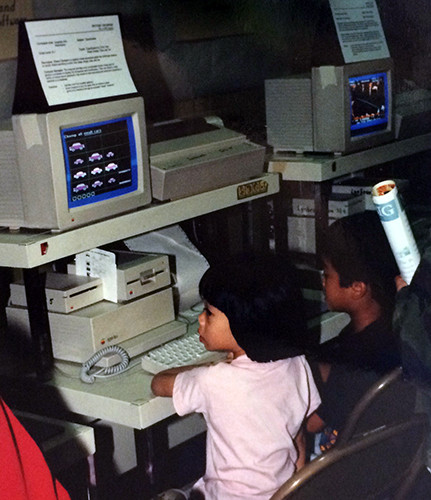 Color photograph of two students sitting at a computer work table. A keyboard, hard drive, and disk drive are visible on the table in front of the children. A CRT monitor and dot matrix printer are on a shelf just above the other computer components. The children appear to be engaged in playing an educational game of some sort.