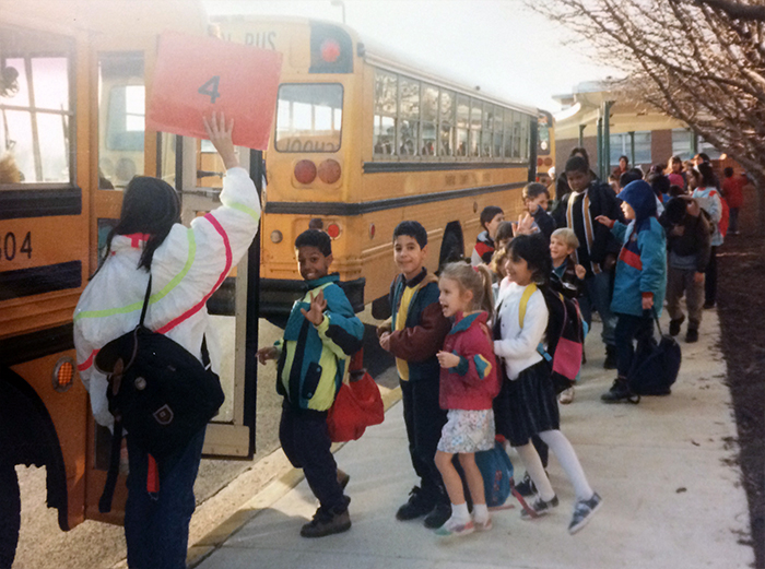 Color photograph of students lining up to board a school bus. An adult in the foreground is holding up an orange sign with the number four printed on it. Three school buses can be seen parked along the sidewalk on the south side of the school.