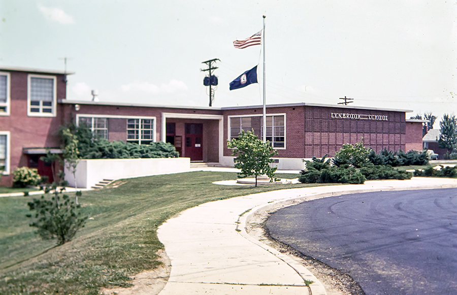 Color 35 millimeter slide photograph of the front exterior of Lynbrook Elementary School. The picture is believed to date from the late 1970s or early 1980s. The original main entrance of the building is shown. The windows have white trim and the doors are painted red.   