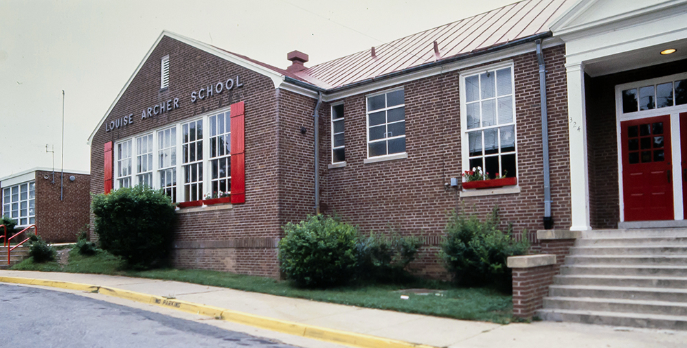 Color photograph of the front exterior of Louise Archer Elementary School. The oldest portion of the building, dating to the 1940s, is shown. It is a brick building with a peaked, tin-roof. The school has white window frames, red shutters, and red doors. 