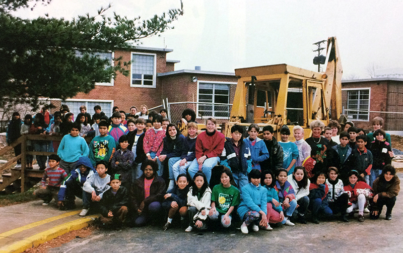 Color photograph of approximately 60 students and two teachers standing in the driveway loop in front of Lynbrook Elementary School. Behind them, the main entrance to the building has been fenced off and the sidewalk has been removed. A large backhoe is parked behind the children.