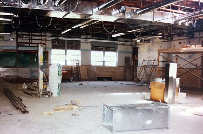 Photograph of a classroom interior during the renovation. The ceiling tiles and light fixtures have been removed and new heating and cooling duct work is being put in place. Scaffolding is visible in two locations, and a large section of metal duct work is visible on the floor. This may be the original library space being converted to classroom use.
