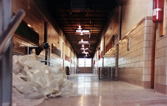Photograph of the second floor hallway. The ceiling tiles have been removed. A plastic covering of some sort is balled up and pushed against the wall on the left side of the picture. An industrial box fan can be seen about halfway down the hallway on the right. New tiles have been added along the walls and floor of the hallway.