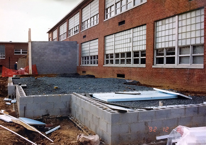 Photograph of the rear of the two-story classroom wing of the building where the new library is being constructed. The foundation of the new library is in place, but the old exterior wall is not yet demolished. The cinderblock wall in the foreground is about three feet high and the space between this new wall and the original exterior is filled with gravel. A section of cinderblock wall reaching up to the height of the second floor is visible in the distance.