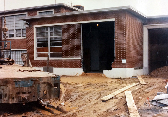 Photograph of a section of the pod classroom wing where part of an exterior wall has been removed. Some construction equipment and supplies are in the foreground. The ground is very muddy and water is pooling in the ruts caused by the tires of construction equipment.