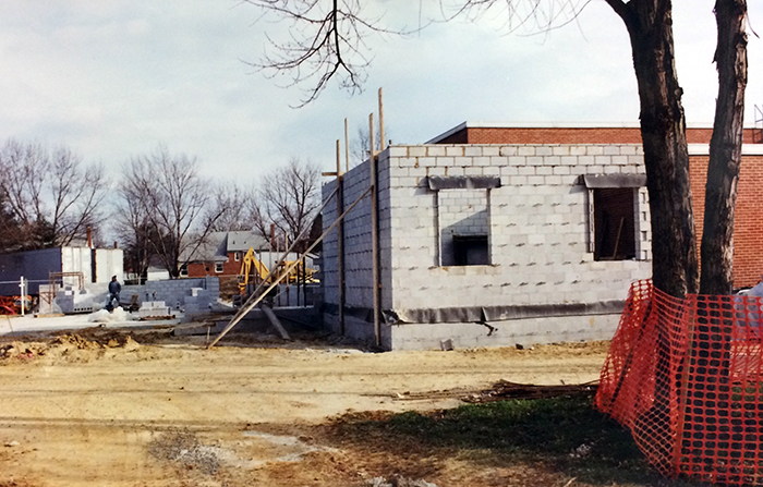 Photograph showing the progress of the construction of the new itinerant office and the SACC classrooms. The cinderblock walls for the office are in place, and the concrete foundation of the SACC rooms have been poured. A construction worker can be seen in the distance, standing next to a section of cinderblock wall being erected on the far side of the SACC classroom space.