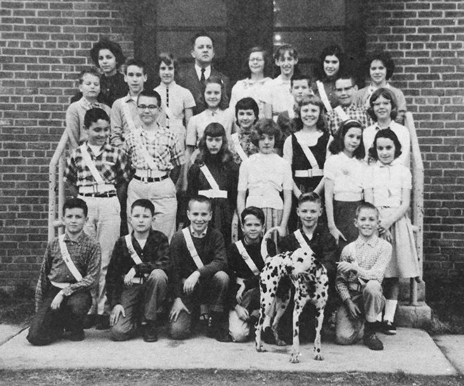 Black and white photograph from the 1960 to 1961 Memory Book showing Lynbrook's Safety Patrol. 27 children and the teacher sponsor are shown. Standing in front of the first row of students is a Dalmatian dog that appears to be wagging its tail. The Safety Patrol members are wearing distinct white belts. 