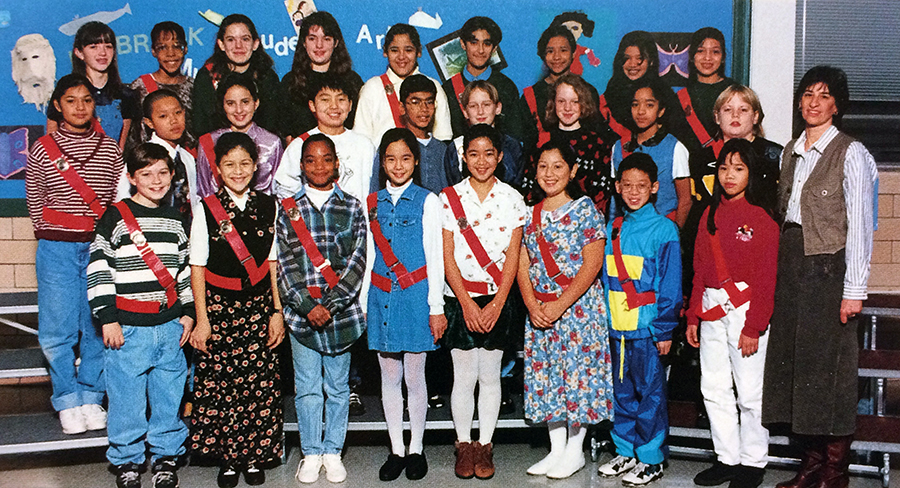 Color photograph from Lynbrook's 1996 to 1997 yearbook showing Lynbrook's Safety Patrol. 26 children and the teacher sponsor are shown. The children are standing on risers in the cafeteria and are arranged in three rows with their teacher on the far right. The children are wearing distinct red bets. 