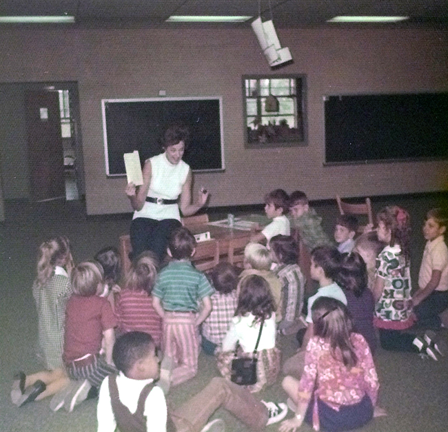 Color photograph of a teacher delivering  a lesson to students in the pod. The pod is a large open space surrounded by classrooms on four sides. The teacher is at center, sitting on a table. She is holding up a paper and speaking to the students who are seated on the carpeted floor in front of her. Approximately 19 children are shown, seated with their backs to the camera. Two blackboards are visible on the wall in the distance, behind which is a classroom with windows.  